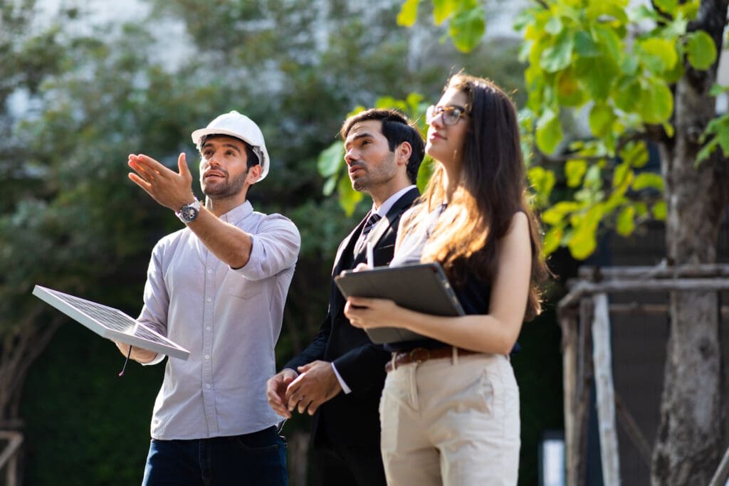 An outdoor team meeting at a construction site with a man in a hard hat explaining details on blueprints, flanked by a man and a woman in business attire, both focused on the project ahead.