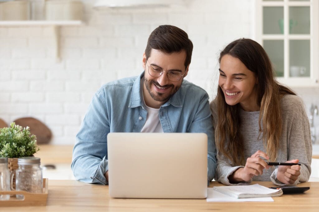 Two smiling homeowners sit at a table, viewing roofing options on a laptop and discussing the potential choices, with a sense of collaboration and shared decision-making.
