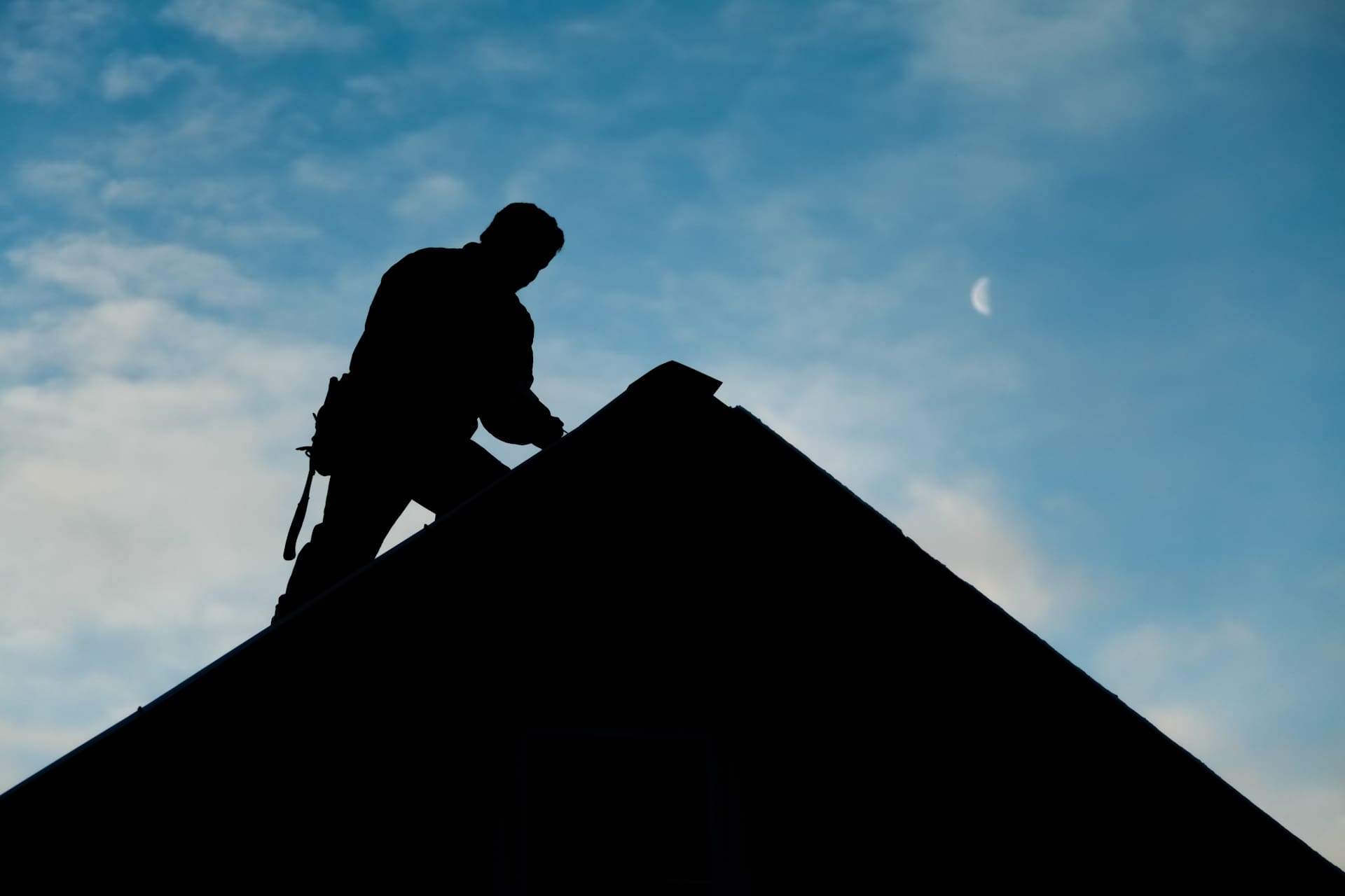 Silhouette of a roofer standing on the ridge of a house against a dusky sky, with a crescent moon visible in the background.